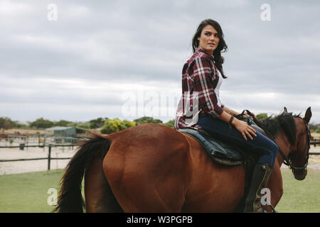 Beautiful young woman riding horse inside corral ranch. Equestrian woman horse riding outdoors. Stock Photo