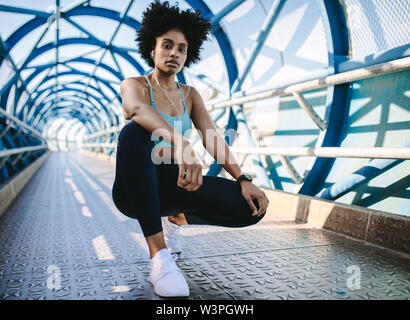 Young woman runner resting after running workout. Female fitness model crouching on a bridge. Stock Photo