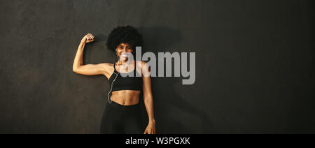 Young fitness woman flexing muscles and smiling. African female model in sportswear showing her muscles against grey background. Stock Photo