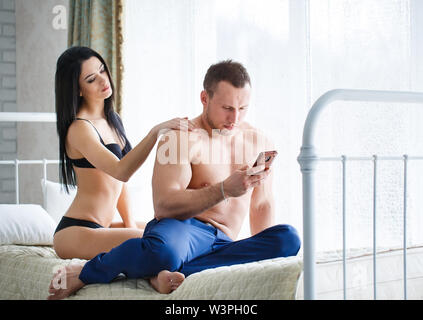 Problems in the family. Angry young woman lying on the bed, against her husband, who plays the game on the phone. Stock Photo
