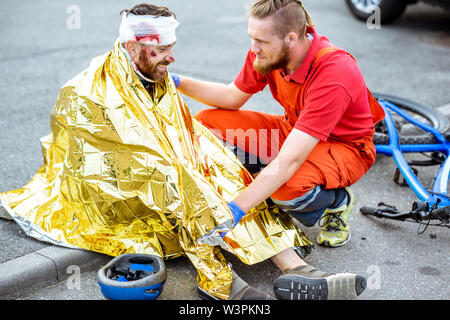 Ambluence worker covering injured man with thermal blanket, providing emergency care after the road accident Stock Photo