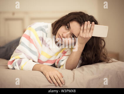 Closeup of young beautiful bored girl on bed using mobile phone. Tired checking smartphone in melancholic mood. Communication internet and technologie Stock Photo