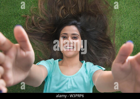 Close up top view of a smiling teenage girl lying on grass with hair spread all around raising her arms Stock Photo