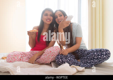 Two smiling young girls sitting on bed at home eating pizza and watching television Stock Photo