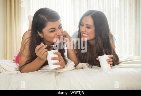 Two smiling teenage girls lying on bed at home holding coffee mugs and spoons and talking with one girl pampering the other Stock Photo