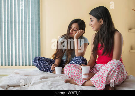 Two smiling teenage girls sitting on bed at home holding coffee mugs and talking Stock Photo