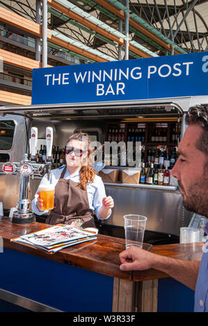 The Winning Post Bar at Royal Ascot racecourse, Berkshire, UK. Barmaid serving a customer with a draught pint of lager. Serving alcohol on sunny day