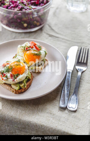Breakfast served of two toasts with avocado, fried eggs with vegetables and herbs on a rustic tablecloth background. View from above. Healthy and nutr Stock Photo