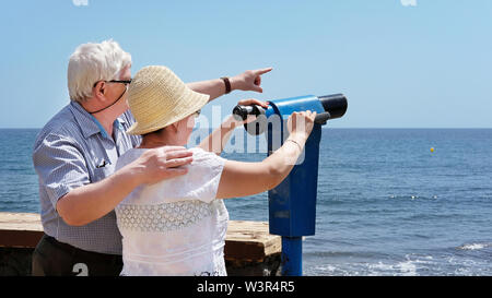 Senior woman wearing hat, holding binocular and man wearing spectacles with hand on woman's shoulder and pointing across the sea in El Medano Tenerife Stock Photo