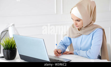 Portrait of young muslim woman student in beige hijab is doing her homework sitting on workplace writing outline using laptop. Stock Photo