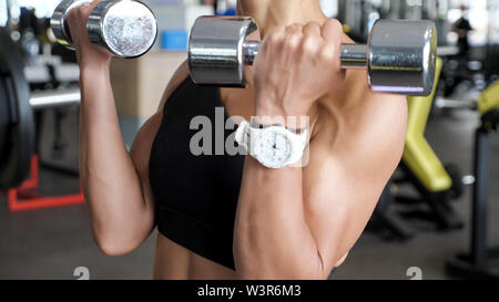 unknown athletic woman is making set of reps exercise for biceps with dumbbells in hands in gym. She is lifting dumbbells, close up Stock Photo