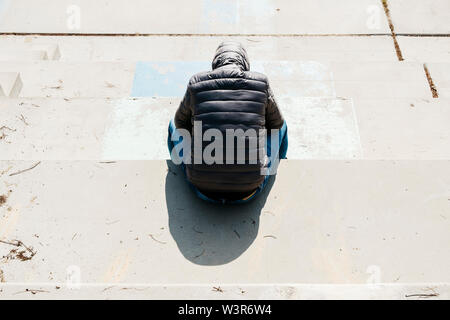 a young caucasian man, seen from behind, wearing jeans and a gray hooded jacket, curled up sitting on an outdoor concrete stairway Stock Photo