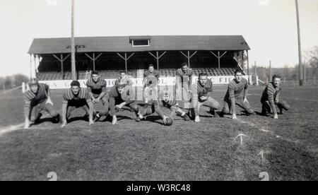 1930s, historical, student players of a college american football team in a group formation outside kneeling on a grass pitch infront of a wooden roofed grandstand, USA. Stock Photo