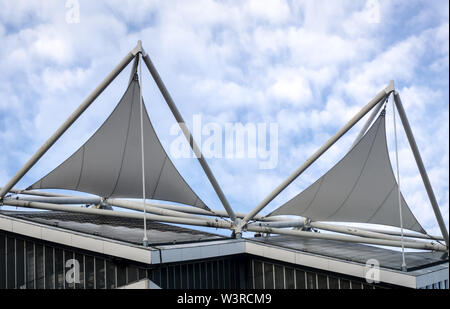 Modern architectural tensile fabric structure featuring white triangular sail canopies supported by a steel mast and cable system against a blue sky. Stock Photo