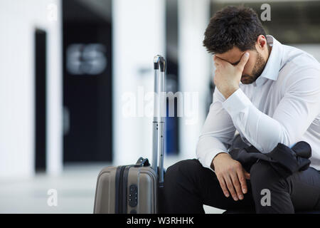 Sad handsome young businessman in white shirt sitting with wheeled luggage in airport and crying because of being late on plane Stock Photo
