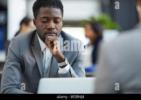 Serious thoughtful handsome young black businessman touching chin while examining report on laptop Stock Photo