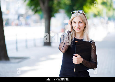 A young beautiful blonde girl wearing a classic black dress holds a folder of papers in her hands. Business clothing style. The girl is working Stock Photo