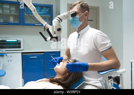 handsome dentist in medical mask examining patient woman laying on the dental chair using professional microscope in clinic Stock Photo