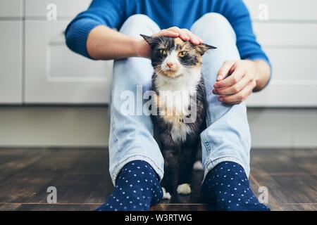 Domestic life with pet. Young man siiting on floor in home kitchen and stroking his cute cat. Stock Photo
