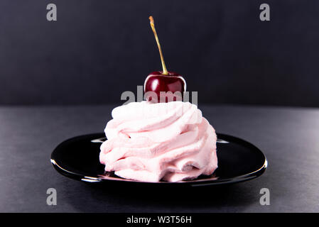 Sweet pink cake with a single cherry on top in a black saucer against a dark background. Pink cherry marshmallows. Red cherry. Stock Photo
