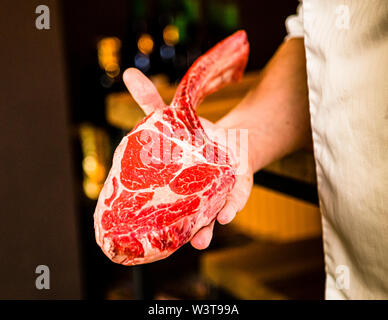 A hand holds a raw tomahawk steak with clearly visible marbling at the Feinkost Faber steakhouse in Bad Kissingen, Germany. The Irish dry-aged cut weighs around one kilogram depending on how it is trimmed and serves two to three people. Stock Photo