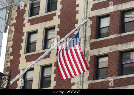 Hanging off the side of one building with brick facade in background, stars and stripes on the flag of America waving in summer. Stock Photo