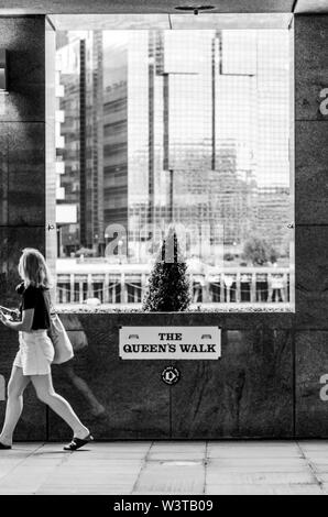 Black and white street photo of a young girl walking on The Queen's Walk promenade in London and passing by the large window with a small tree and an Stock Photo