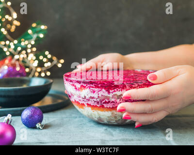 Woman hands with layered salad herring under a fur coat on festive table. Traditional russian salad with herring and vegetables in glass bowl. Copy space for text. Stock Photo