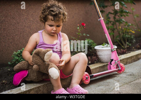 Sweet little girl with curly hair is sitting and resting near her pink scooter in the courtyard, holding a teddy bear Stock Photo