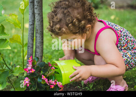 Sweet little girl with curly hair watering colorful flowers and a tree in the beautiful garden. Children ecology concept Stock Photo