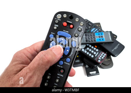 Horizontal close-up shot of a woman’s hand holding a remote control with blue, gray, and red buttons.  There is a pile of old remote controls in the b Stock Photo