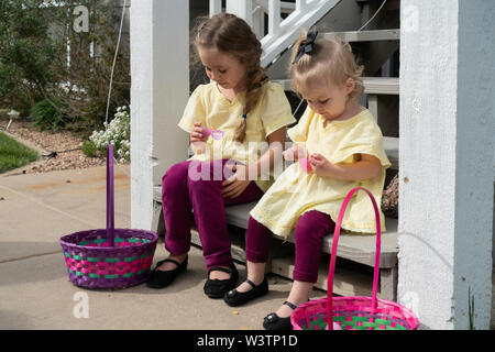 Two Caucasian girls, 2 years old and five years old, sit on stairs and open plastic Easter eggs and eat treats from them. USA Stock Photo