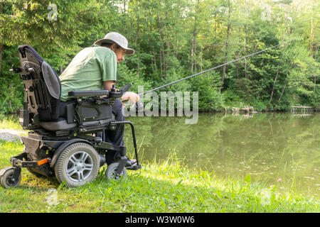 Happy man in a electric wheelchair waiting to catch a fish at the beautiful pond in natue on a sunny day Stock Photo