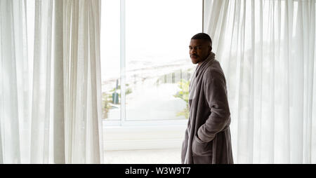 Man in bathrobe looking at camera while standing near window in bedroom Stock Photo