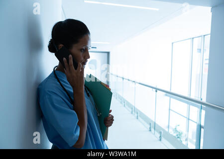 Female surgeon talking on mobile phone while leaning against wall in the corridor of hospital Stock Photo