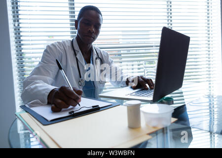 Male doctor writing on a clipboard while using laptop at desk Stock Photo