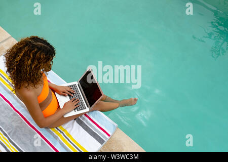 Woman using laptop while sitting at the edge of swimming pool Stock Photo