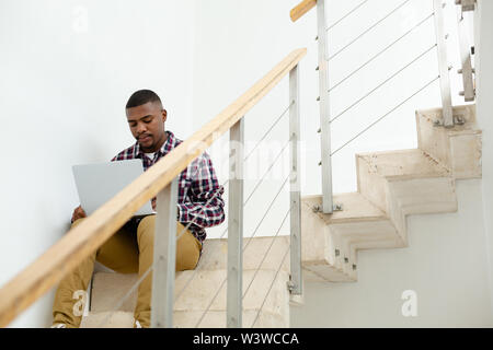 Man working on laptop while sitting on stairs in a comfortable home Stock Photo