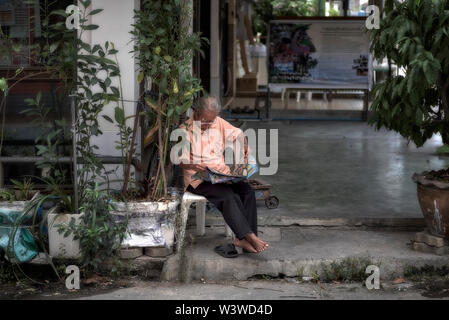 Man reading the newspaper on the pavement outside of his home. Thailand Southeast Asia Stock Photo