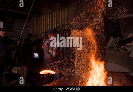 two real brutal traditional blacksmith workers working together with a red hot iron using mechanical hammer at workshop  Portrait of a profession Stock Photo