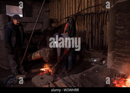two real brutal traditional blacksmith workers working together with a red hot iron using mechanical hammer at workshop  Portrait of a profession Stock Photo