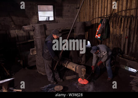 two real brutal traditional blacksmith workers working together with a red hot iron using mechanical hammer at workshop  Portrait of a profession Stock Photo
