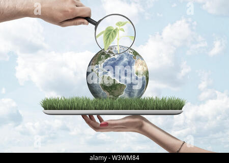 Magnifying glass in man's hand zooming at green sprout on small planet Earth lying on screen of digital tablet that is resting on woman's palm. Stock Photo