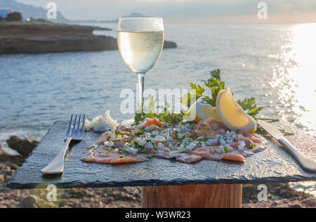 Tasty lunch in front of the sea - Smoked salmon with chopped onion, celery, lemon slices and a glass of wine - close-up Stock Photo