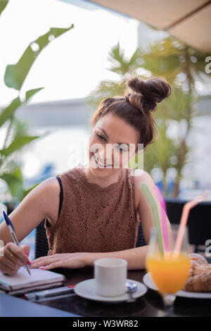 Adorable young woman taking notes with orange juice bread and espresso coffee buns on the table - green plants background Stock Photo