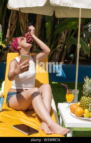 Beautiful girl eating grapes on the terrace while taking a photo with her cell phone - Exotic fruits and orange juice on the table - Banana trees in t Stock Photo