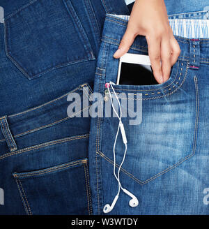 hand holding white smartphone with a blank black screen and headphones is in the front pocket of blue jeans, close up Stock Photo