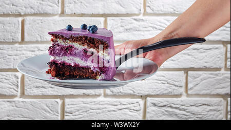 a woman hand holding a portion of blueberry and blackberry chocolate sponge cake layered with cream cheese on a plate with brick wall at the backgroun Stock Photo