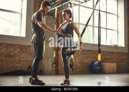 My personal trainer. Confident young man is showing slim athletic woman how to train legs with Trx fitness straps while training at gym. TRX Training. Exercising together. Active lifestyle Stock Photo