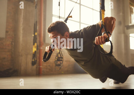Working hard. Side view of young and strong athletic man in sportswear doing push ups with trx fitness straps in the gym. Fitness. TRX Training. Workout concept Stock Photo